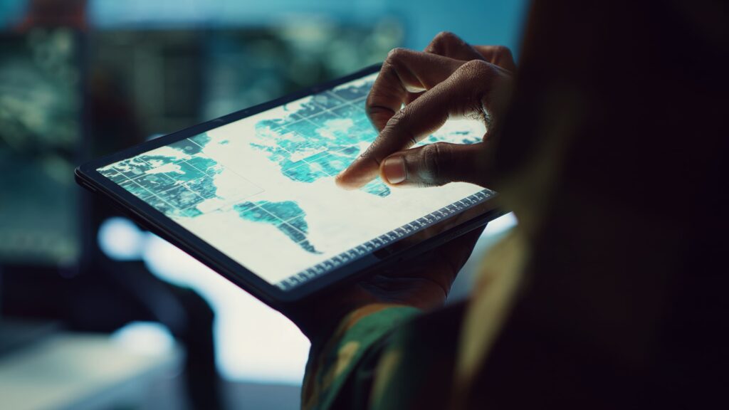 Close-up of a tactical logistics operator's hand using a rugged tablet to navigate a detailed digital world map. Illustrates global supply chain visibility and control provided by KCE Logistics.