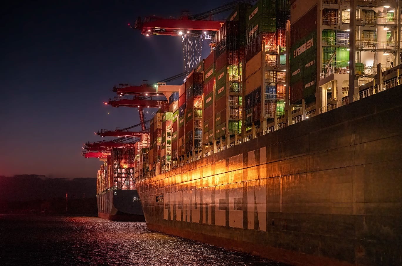 Nighttime view of large Evergreen container ships at a busy terminal, representing the high-stakes nature of maritime logistics during Middle East supply chain disruptions.